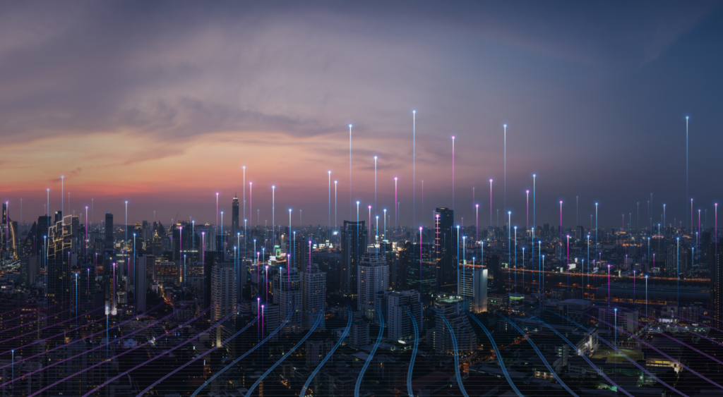 Imagem área de uma cidade durante a noite, com diversos prédios e edifícios comerciais. A cidade é atravessada por diversas faixas azuis de energia na horizontal e na vertical, simbolizando a conexão 5G.