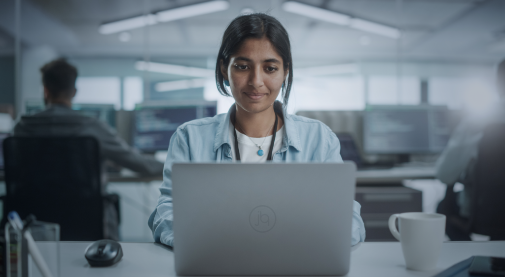 Mulher negra sentada diante de um notebook em um escritório. Ela está vestindo uma camiseta branca, uma camisa social azul clara e um pingente enquanto executa seu trabalho. Isso simboliza o trabalho do programador.